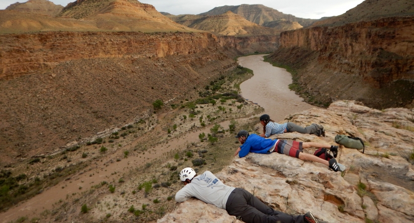 Three people lay on a cliff above a river, looking down. 